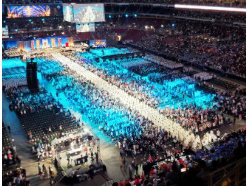 A large crowd illuminated by blue lighting gathers in a ceremonial formation at a religious event.