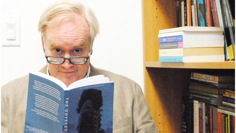 A man with glasses reading "The Odyssey" book, with bookshelves visible in the background.