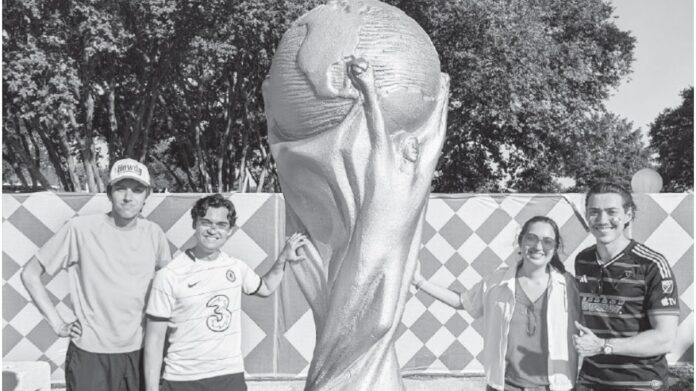 summer-of-dallas-sports-1 People posing in front of a large World Cup trophy replica at an outdoor fan fest with a diamond-patterned backdrop and trees in the background.