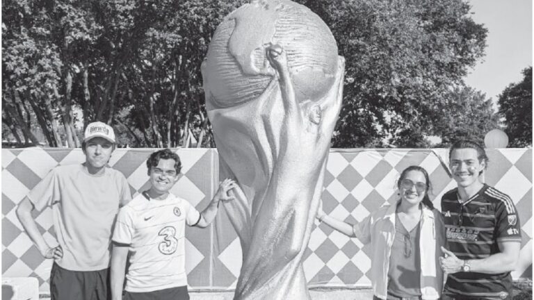 People posing in front of a large World Cup trophy replica at an outdoor fan fest with a diamond-patterned backdrop and trees in the background.
